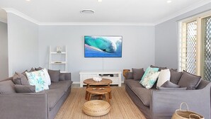 A modern living room with two grey sofas, a wooden coffee table, and a flat-screen TV mounted on the wall displaying a surfing scene.