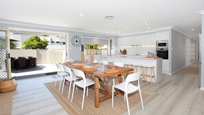 A modern kitchen with white cabinetry, a central island, and a dining area with a wooden table and white chairs. - Beautiful Beach House in the Heart of Town (Coolum Beach)