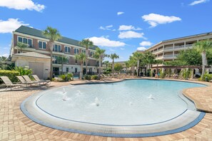 A swimming pool with a fountain, surrounded by lounge chairs and palm trees, with buildings in the background.