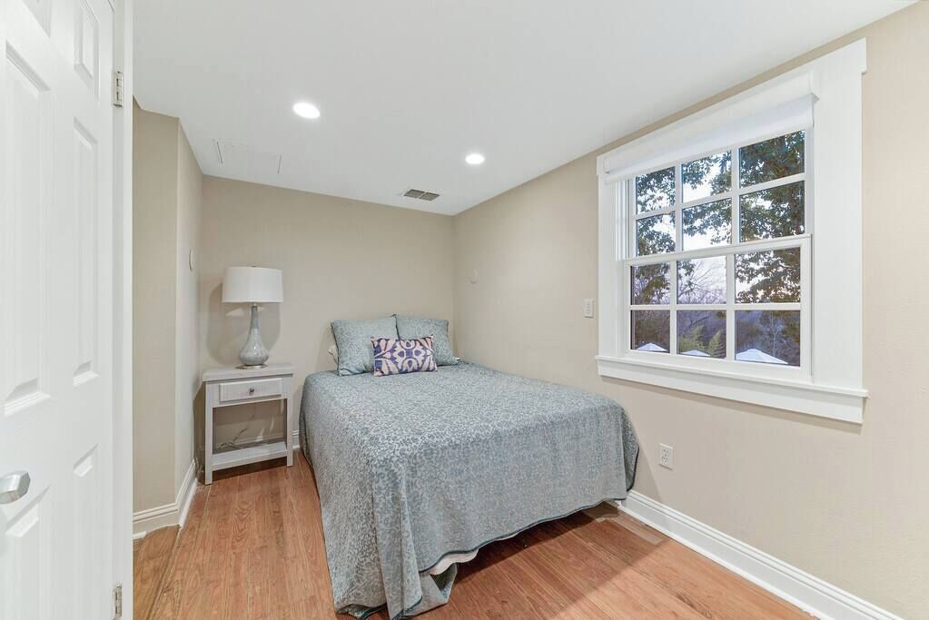 A bedroom with a bed, nightstand, lamp, and a window with a view of trees.