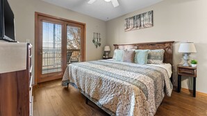 Une chambre avec un grand lit, un ventilateur de plafond, une tête de lit en bois, deux tables de chevet avec des lampes, un téléviseur fixé au mur et une fenêtre avec des stores.