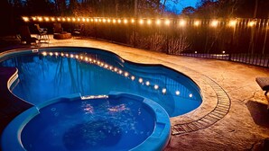 Une piscine avec un jacuzzi, entourée d’une terrasse en pierre et illuminée de guirlandes lumineuses.