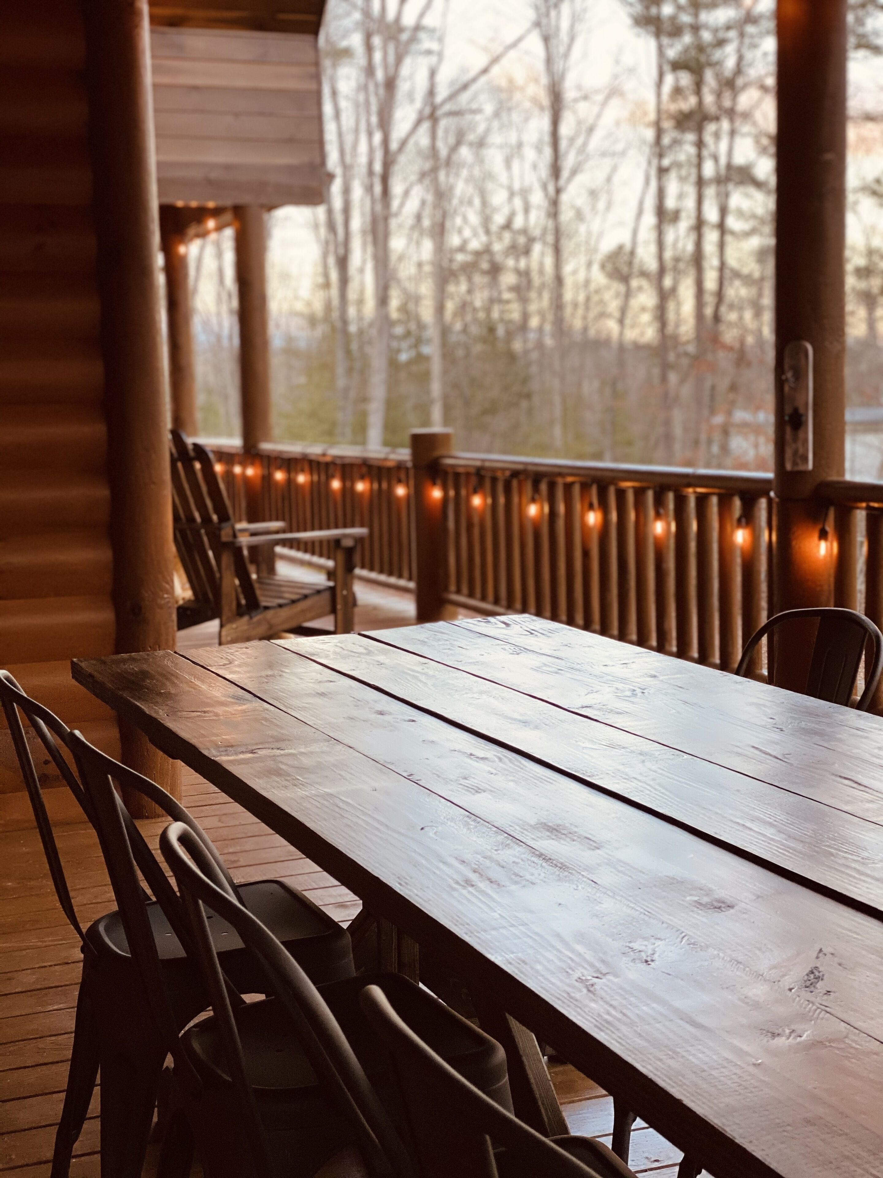 A wooden deck with a table and chairs, string lights, and a view of trees.