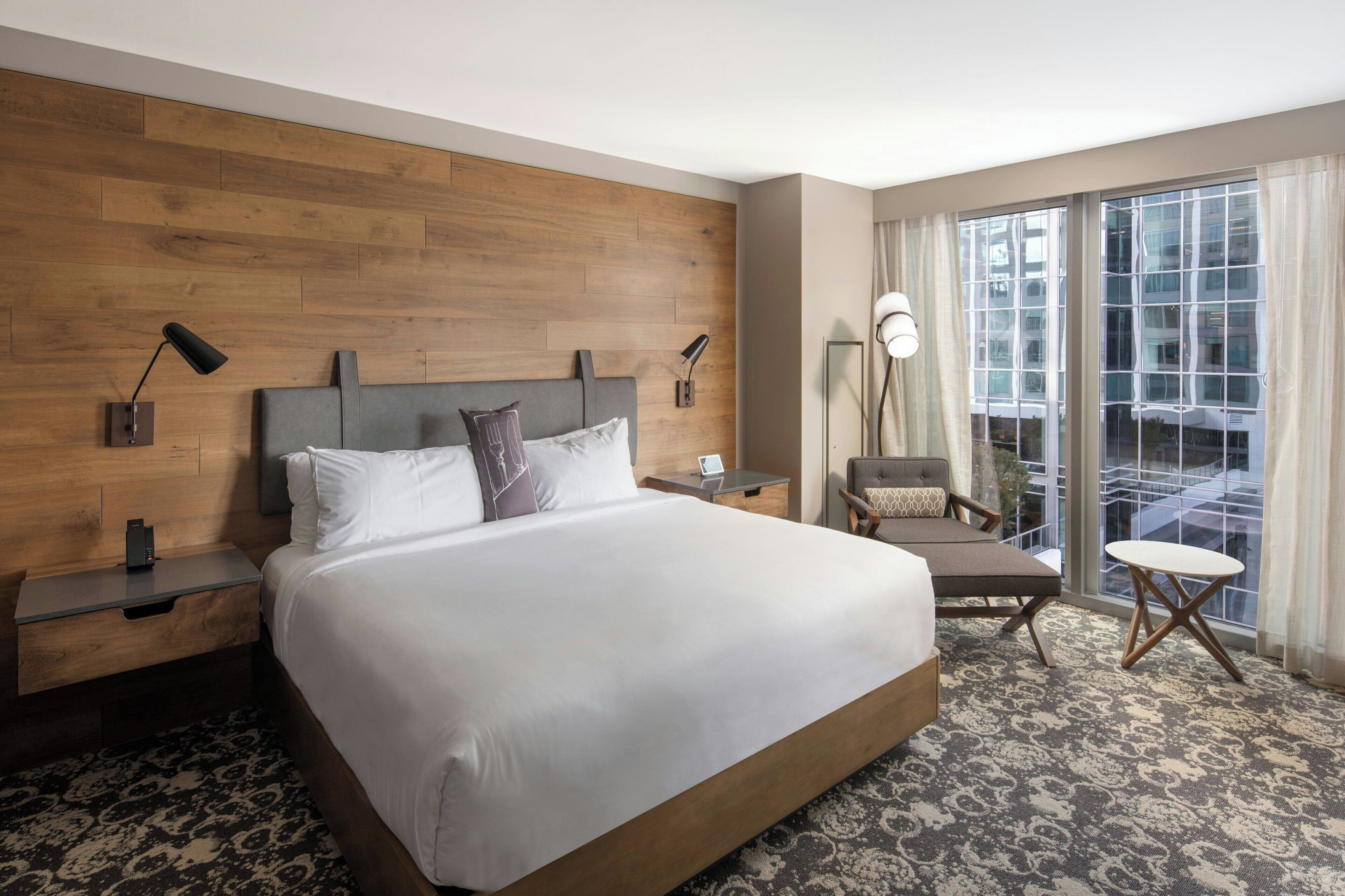 Une chambre d’hôtel avec un grand lit, une table de chevet, un bureau, une chaise et une vue sur la ville grâce à de grandes fenêtres.