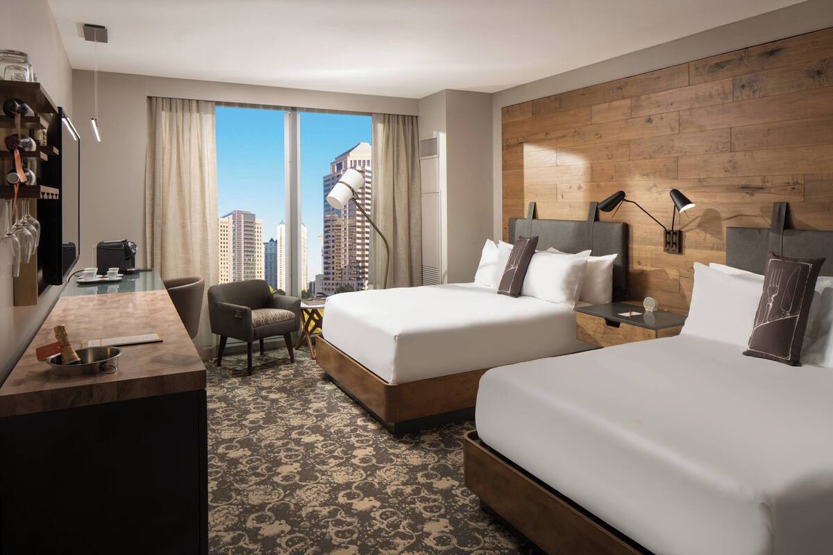 A hotel room with two beds, a desk, a chair, a small table, and a view of the city.