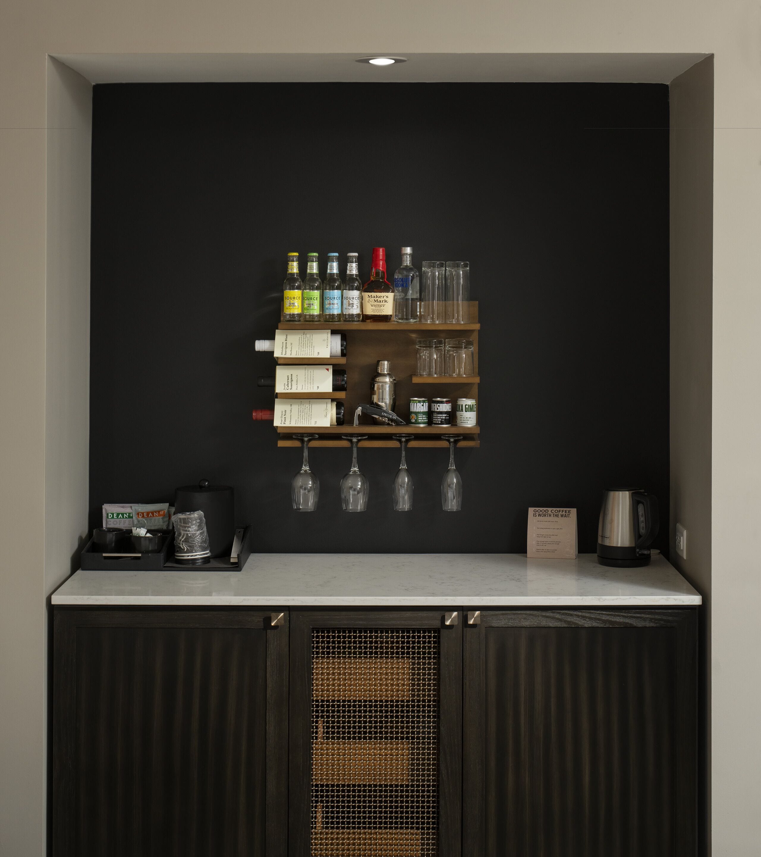 A kitchen with a dark backsplash, a wooden shelf with various bottles and glasses, a coffee machine, and a sign that reads "Good coffee is worth the wait."
