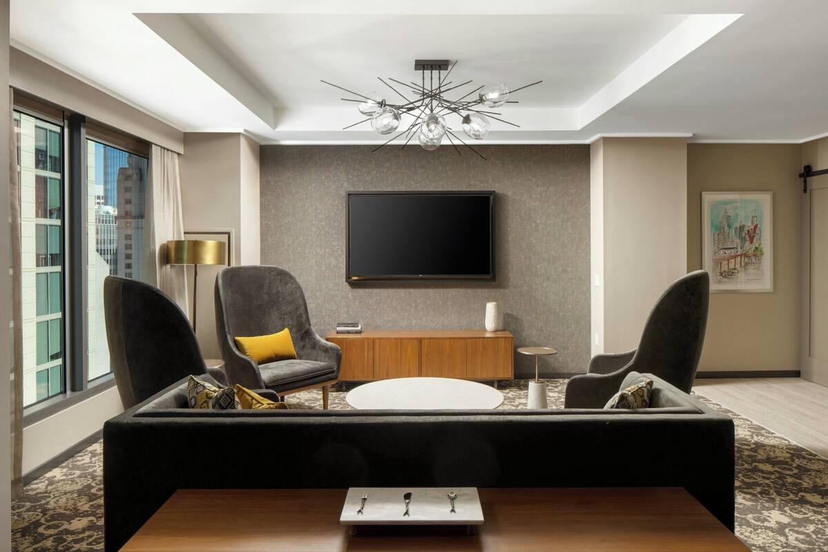 A modern living room with a sofa, two armchairs, a coffee table, a TV mounted on the wall, and a large window with a view of buildings.