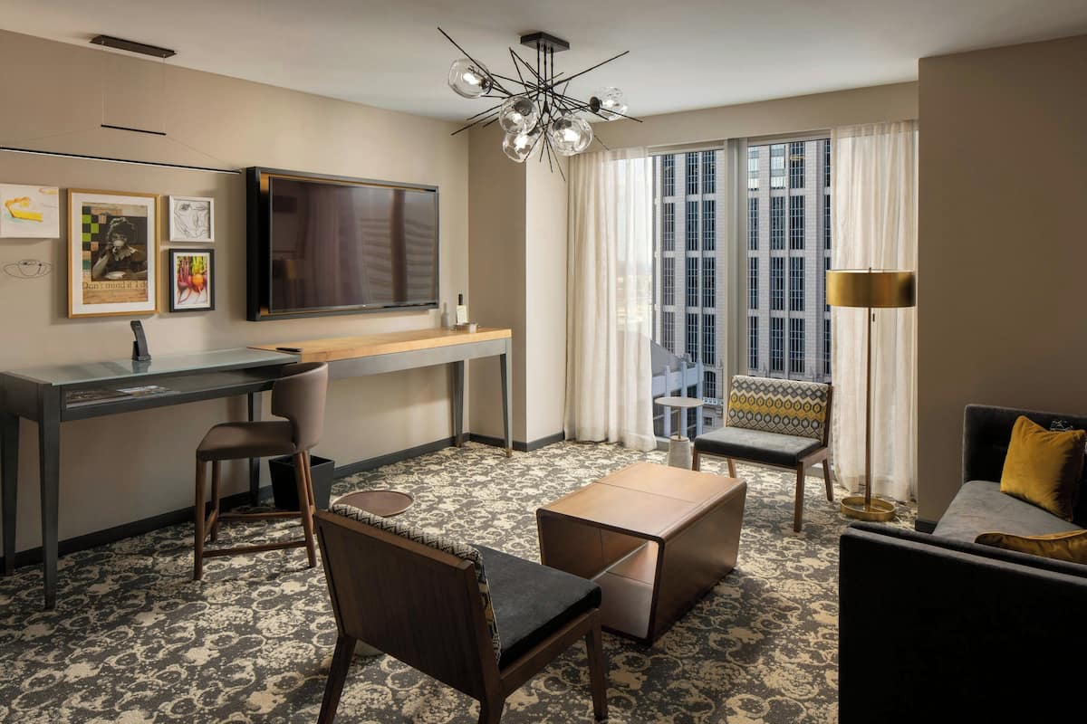 A hotel room with a desk, a television, a sofa, a chair, and a chandelier.