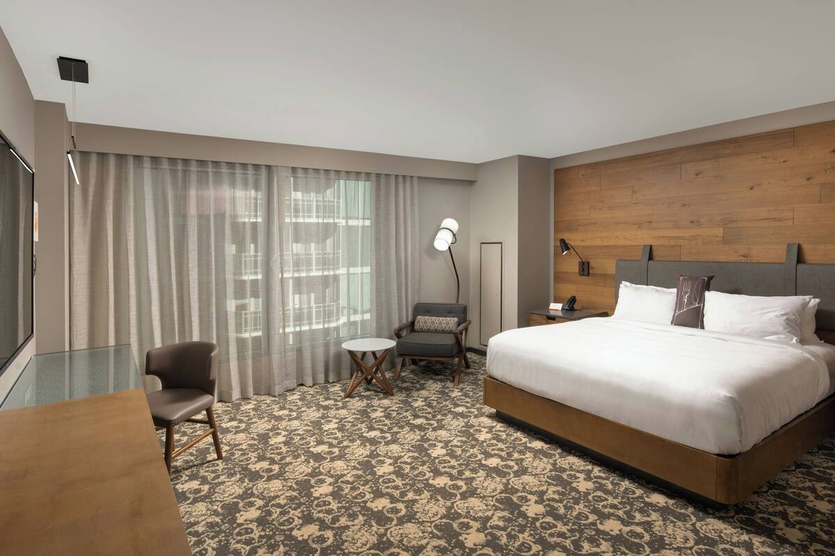 A hotel room with a large bed, a desk, a chair, a small table, and a large window with curtains.