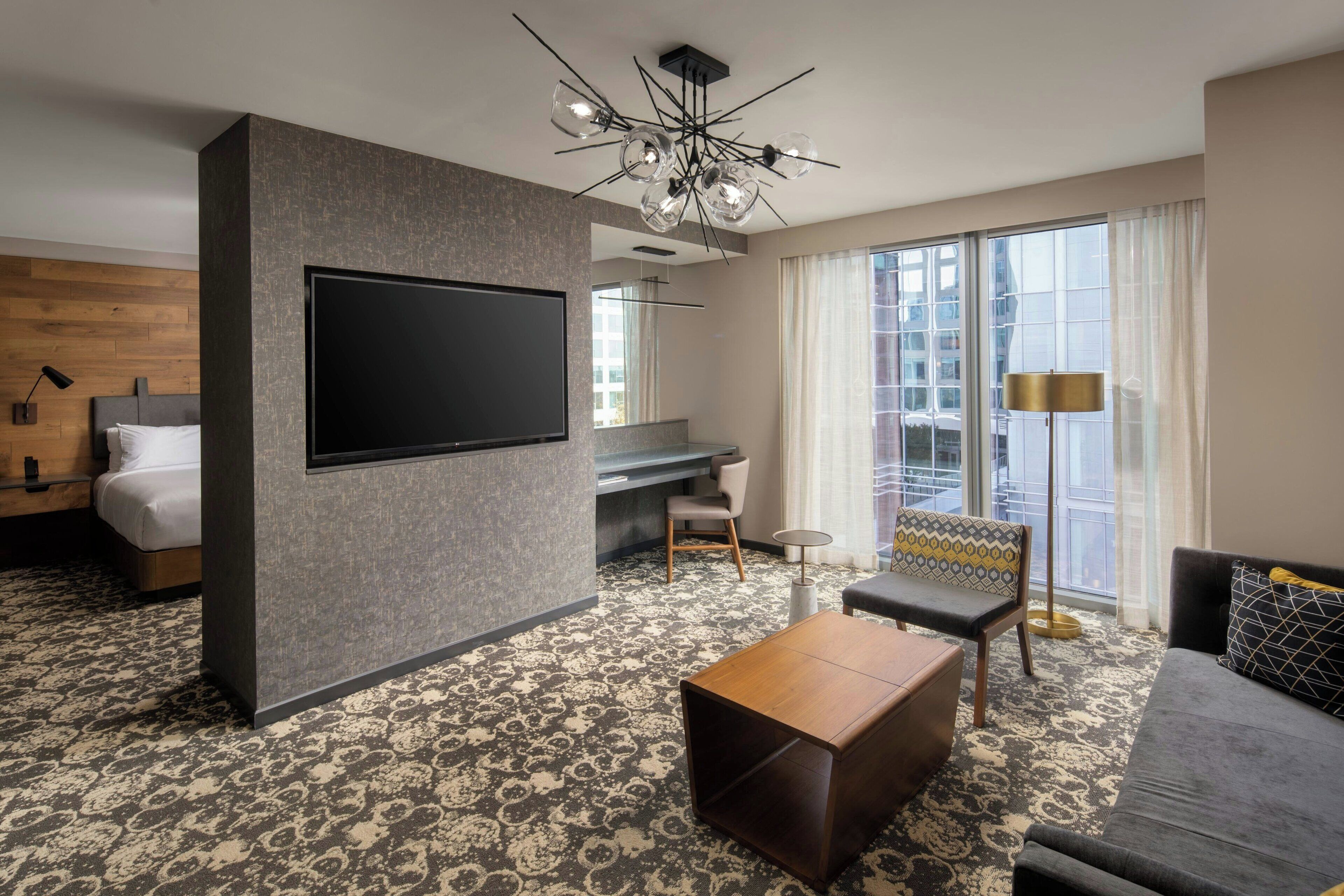 A modern hotel room with a large flat-screen TV, a sofa, a coffee table, a patterned carpet, and a large window with curtains.