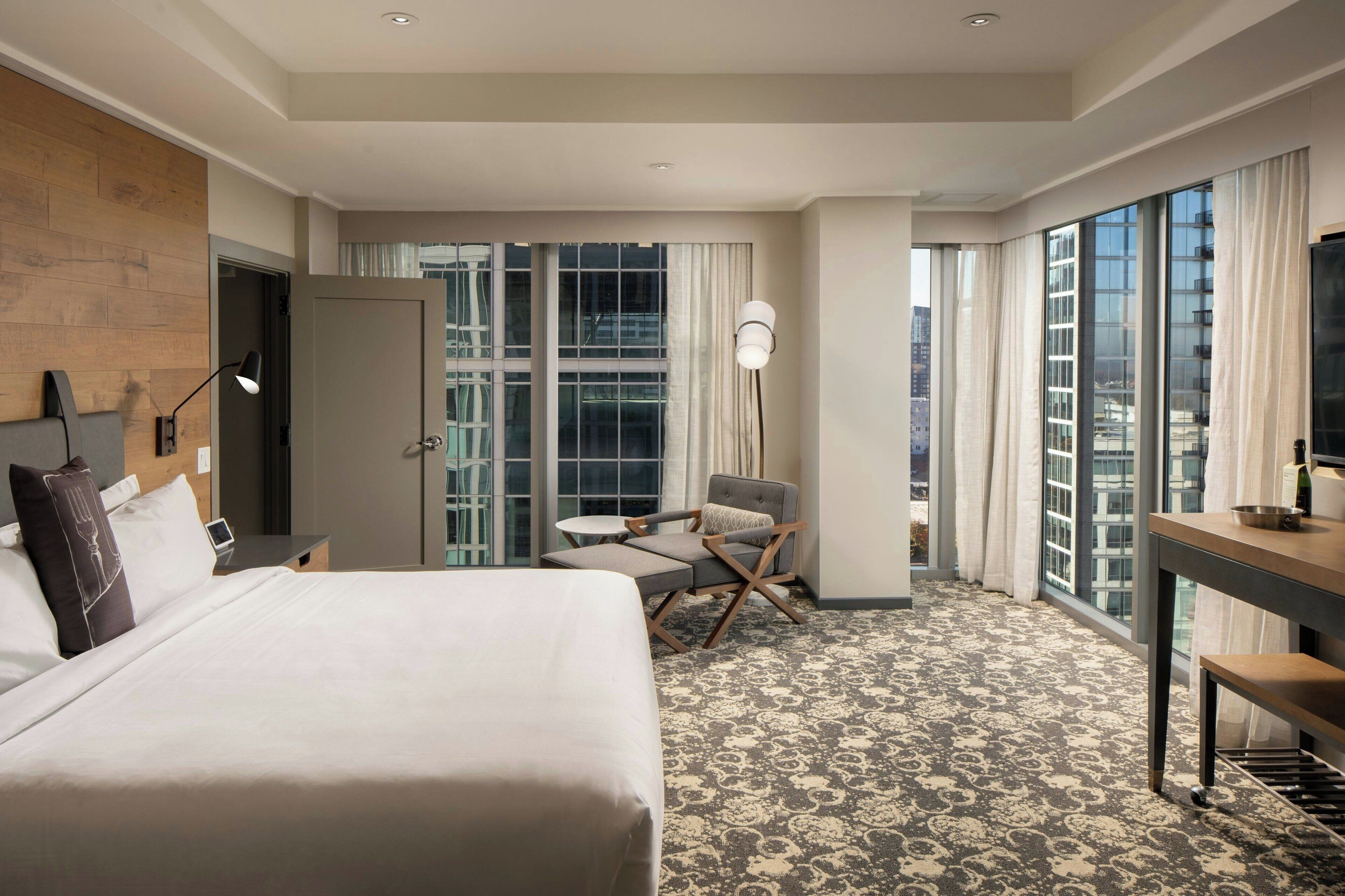 A hotel room with a large bed, a desk, a chair, and a view of the city through large windows.