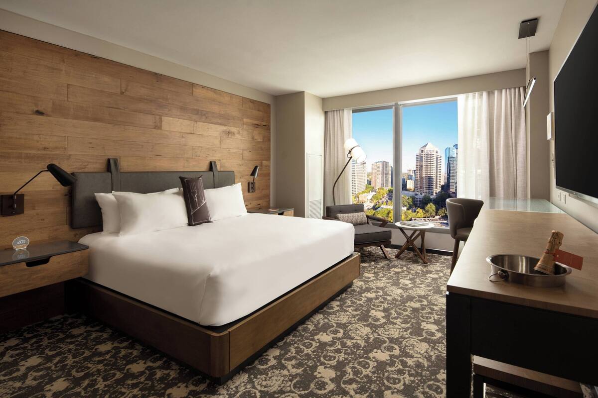 A hotel room with a large bed, a desk, a chair, a small table, a lamp, and a view of the city.