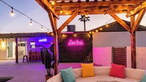 An outdoor seating area with a neon sign reading "Love Oasis," surrounded by string lights and a wooden pergola.