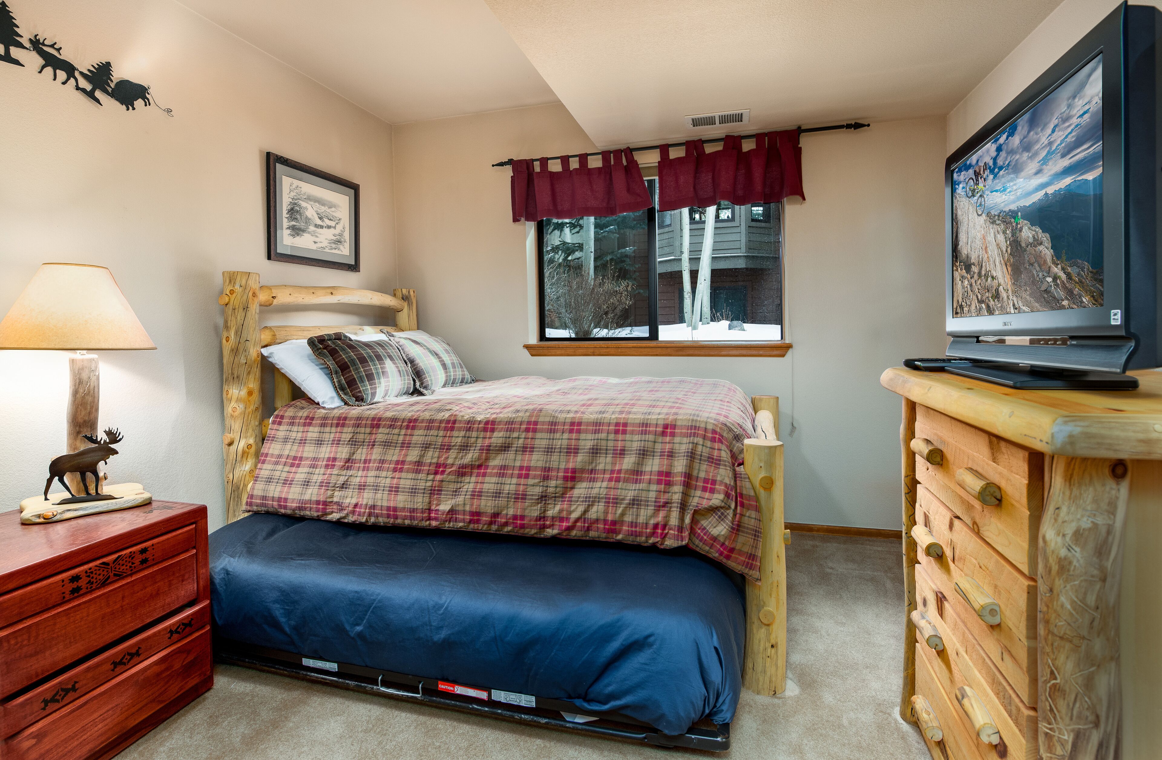 Een slaapkamer met een bed, een dressoir, een televisie, een raam met gordijnen, een lamp en een decoratie aan de muur.