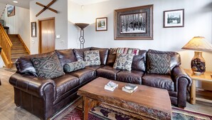 Un salon avec un canapé d’angle en cuir marron, une table basse en bois et un tapis à motifs. On y trouve un escalier avec des rampes en bois et une porte en bois.