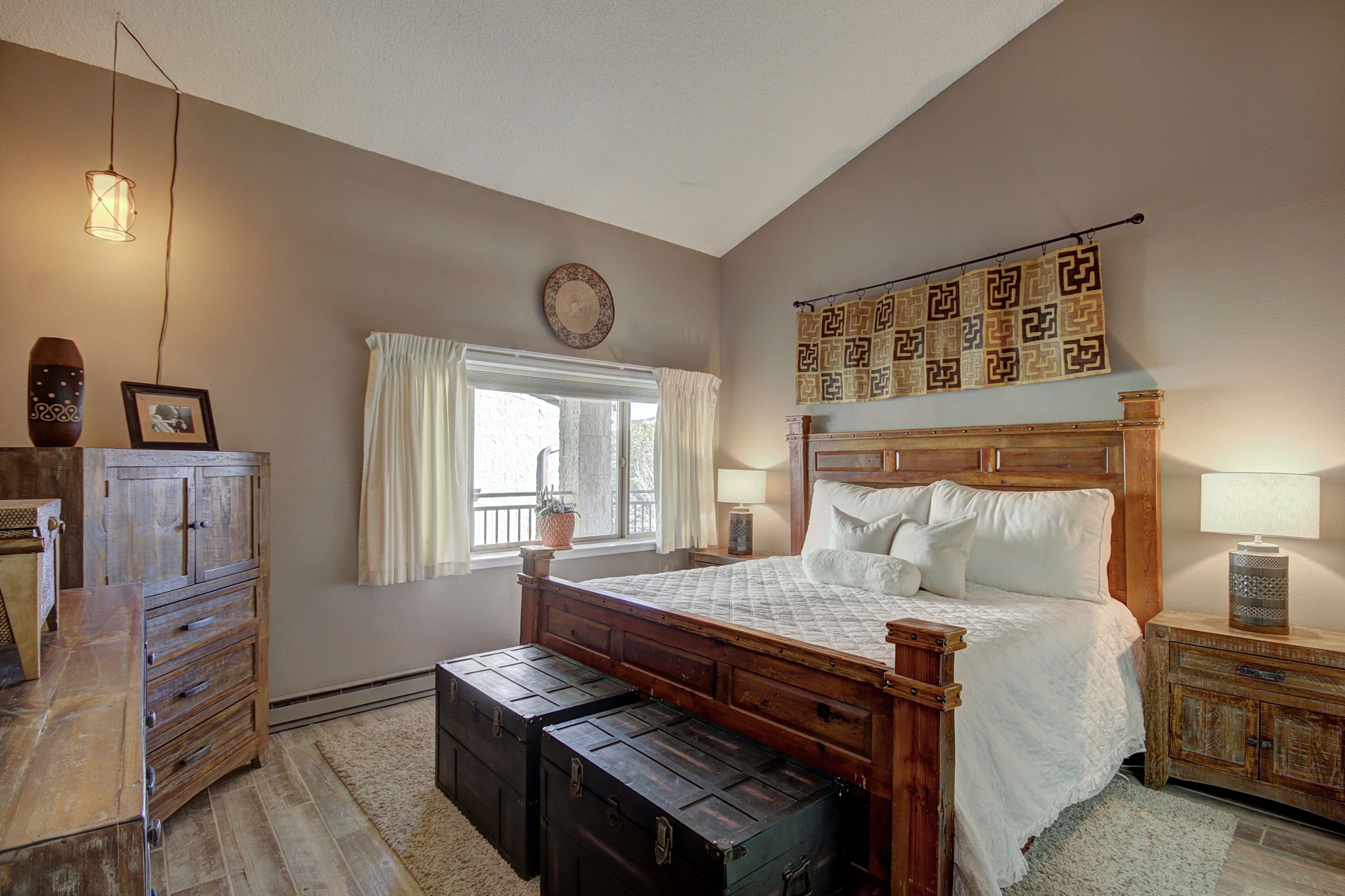 A bedroom with a wooden bed, a dresser, a window with curtains, a wall clock, and a decorative tapestry.