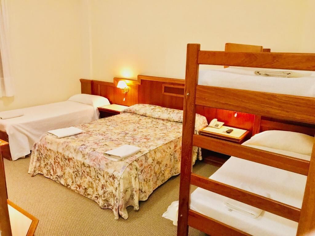 A hotel room with two single beds, a bunk bed, a bedside table with a lamp, and a wall-mounted phone.