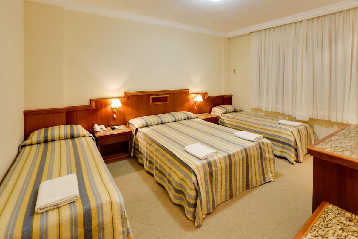 Ein Hotelzimmer mit drei Einzelbetten, jedes mit gestreifter Bettwäsche und einem weißen Handtuch. Es gibt einen Holzkopfbogen, Nachttische mit Lampen und ein großes Fenster mit Vorhängen.