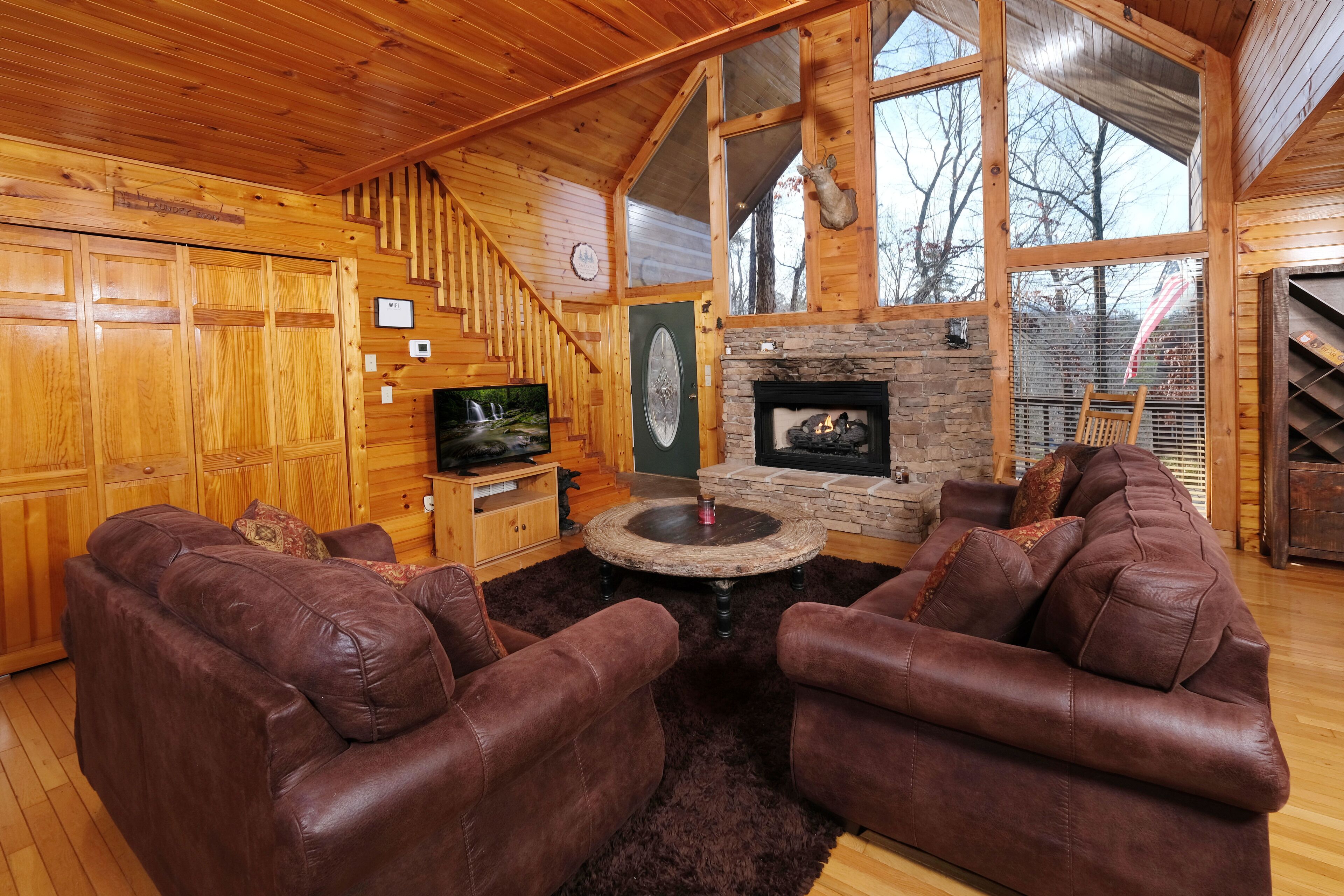 L’intérieur chaleureux d’un chalet avec une cheminée en pierre, des canapés en cuir, un escalier en bois et de grandes fenêtres.