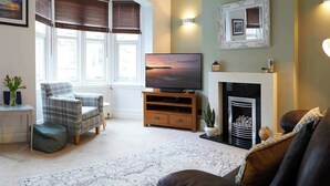 A living room with a fireplace, a television, a plaid armchair, and a view of the outside through large windows. - Fermain Apartment - Spacious cosy duplex apartment - Sleeps 5 (Filey)