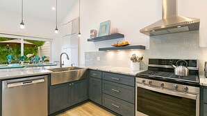 A modern kitchen with dark cabinets, stainless steel appliances, and a large window overlooking a garden. - Gorgeous Oceanview Home! Steps to Beach, Pool & Pickleball (Encinitas)
