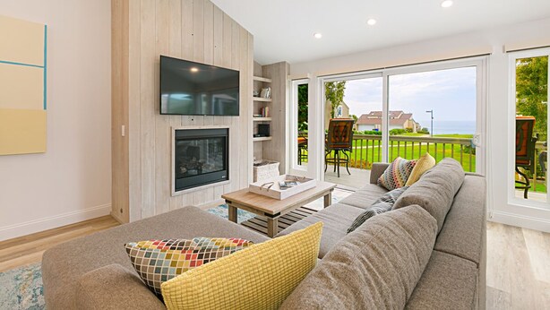 A modern living room with a large sofa, a flat-screen TV mounted on a wooden wall, a fireplace, and a view of the outdoors through large sliding glass doors. - Gorgeous Oceanview Home! Steps to Beach, Pool & Pickleball (Encinitas)