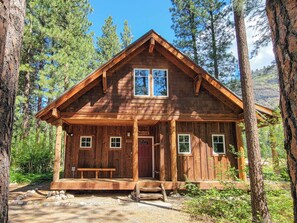 Cozy cabin nestled among tall pine trees, featuring a charming front porch with a bench and windows above.
