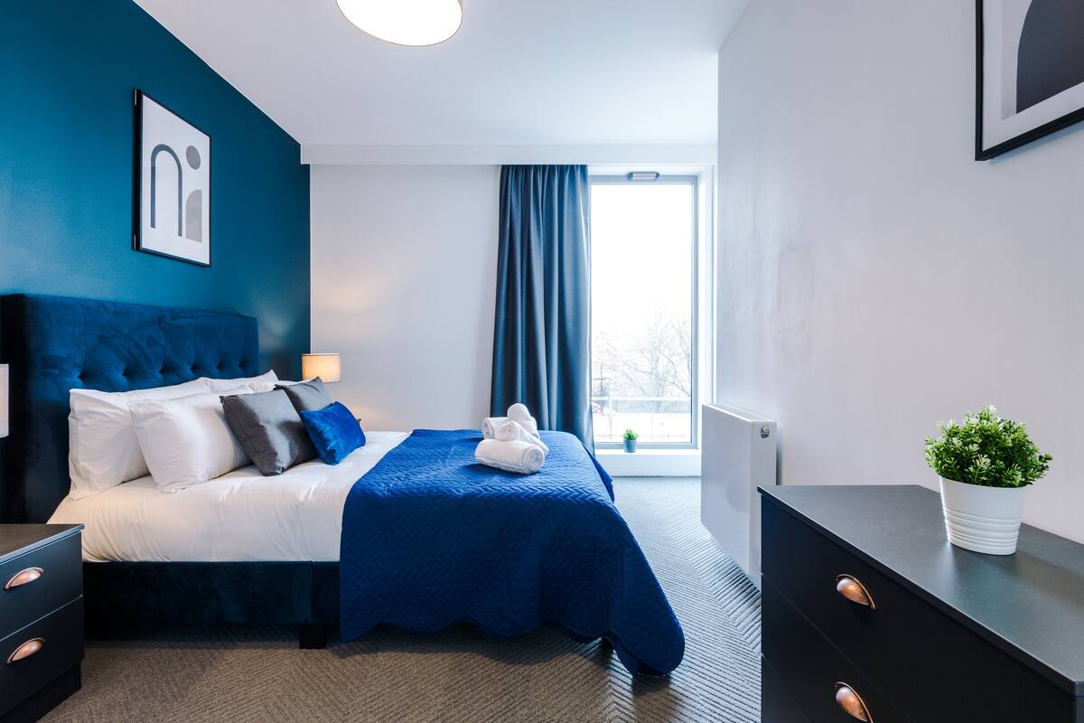 A modern hotel room with a large bed, a nightstand, a dresser, and a window with curtains.
