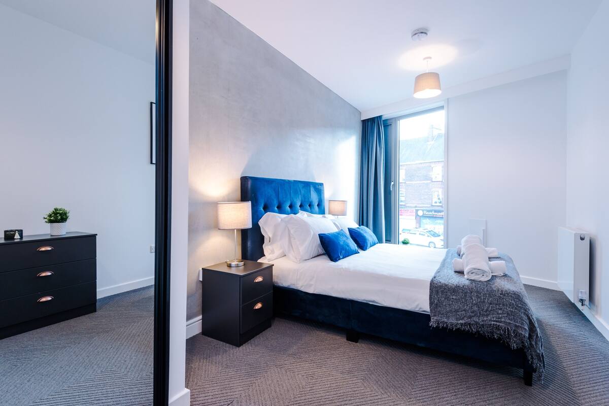 A modern hotel room with a large bed, a nightstand, a dresser, and a view of the city through the window.