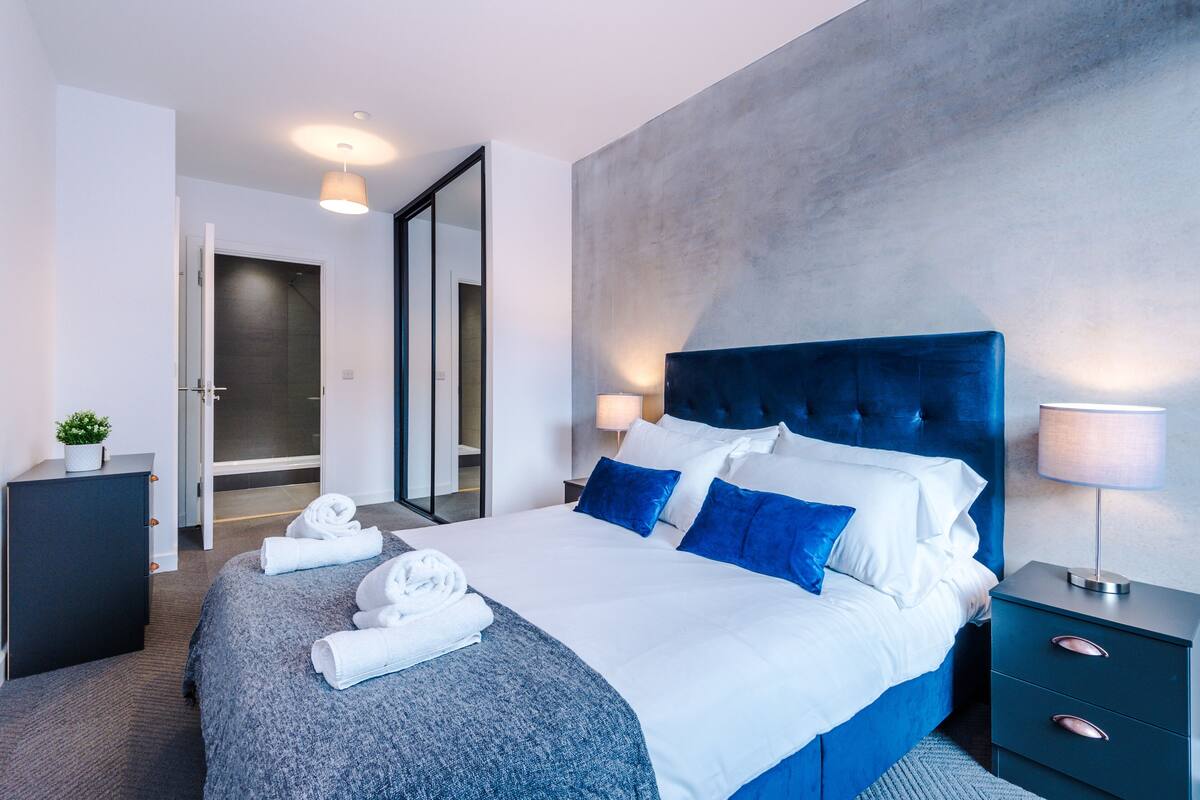 A modern hotel room with a large bed, a dark headboard, and a nightstand with a lamp.