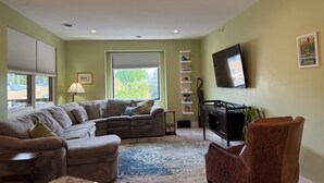 A living room with a sectional sofa, a brown armchair, a TV on the wall, and a window with blinds. - Traverse City, West side of downtown, Sleeps 6, Slabtown area (Traverse City)