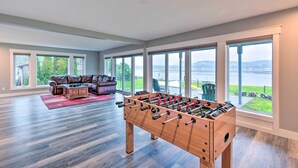 Un salon spacieux avec une table de baby-foot, un canapé en cuir et de grandes fenêtres donnant sur un plan d’eau.
