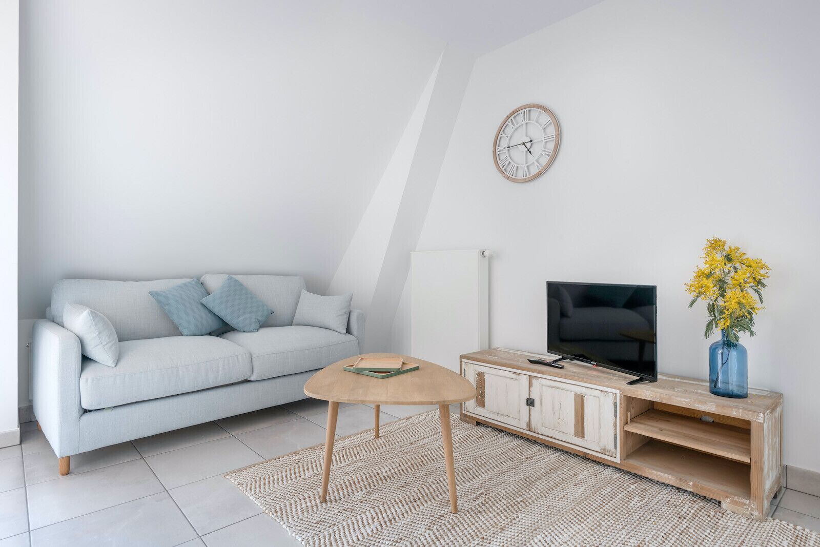 A modern living room with a light grey sofa, a wooden coffee table, a TV on a wooden stand, and a clock on the wall.