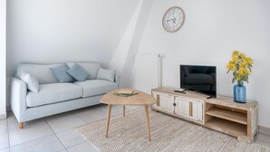 Un saló modern amb un sofà gris clar, una taula de centre de fusta, un televisor sobre un suport de fusta i un rellotge a la paret.