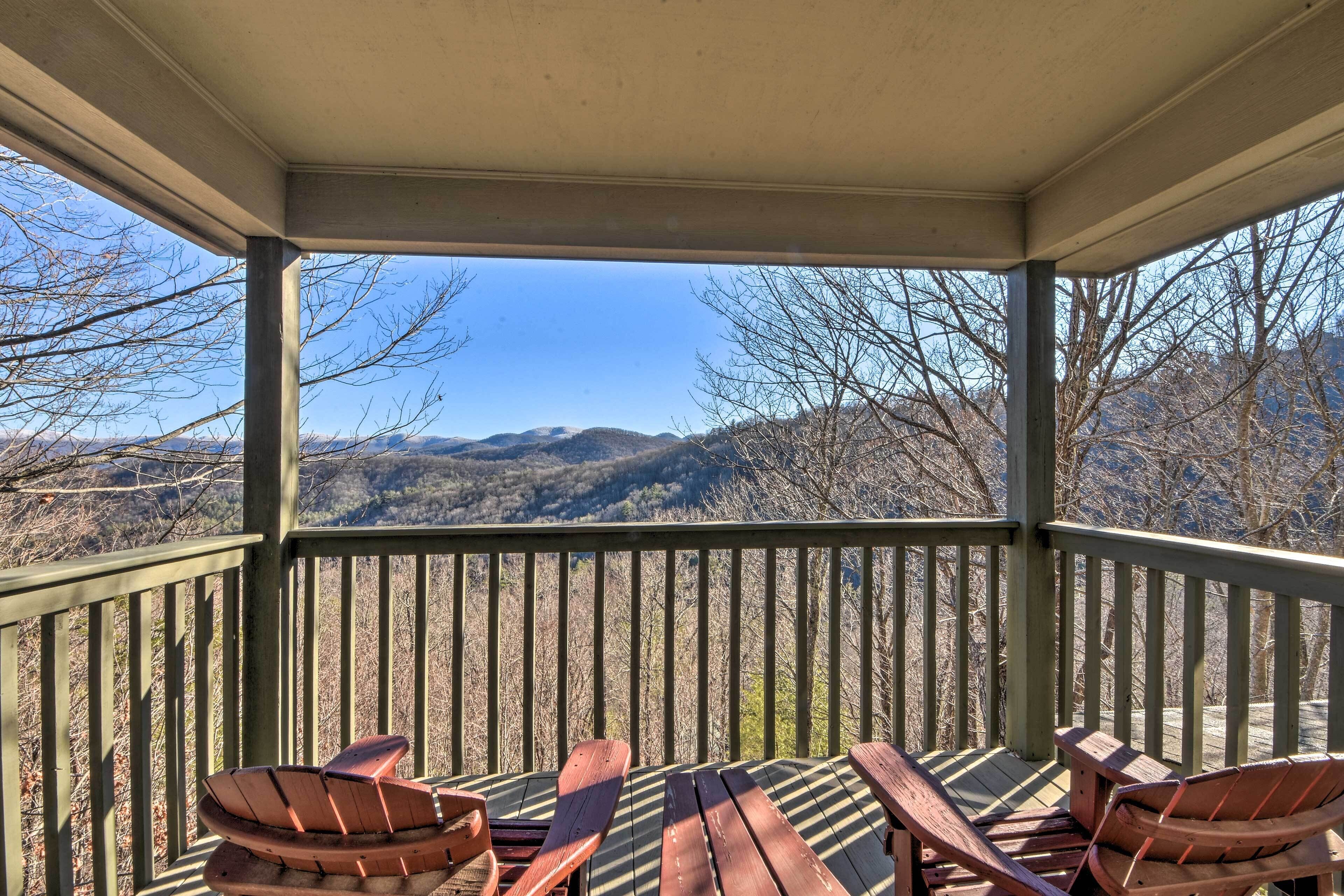 A covered outdoor seating area with wooden chairs and a table, overlooking a mountainous landscape.