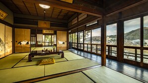 Een traditionele Japanse kamer met tatami vloeren, een lage houten tafel en grote ramen met uitzicht op een stadsbeeld en heuvels.