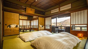 Een traditionele Japanse kamer met tatami vloeren, twee bedden, een bureau en uitzicht op een bergachtig landschap.