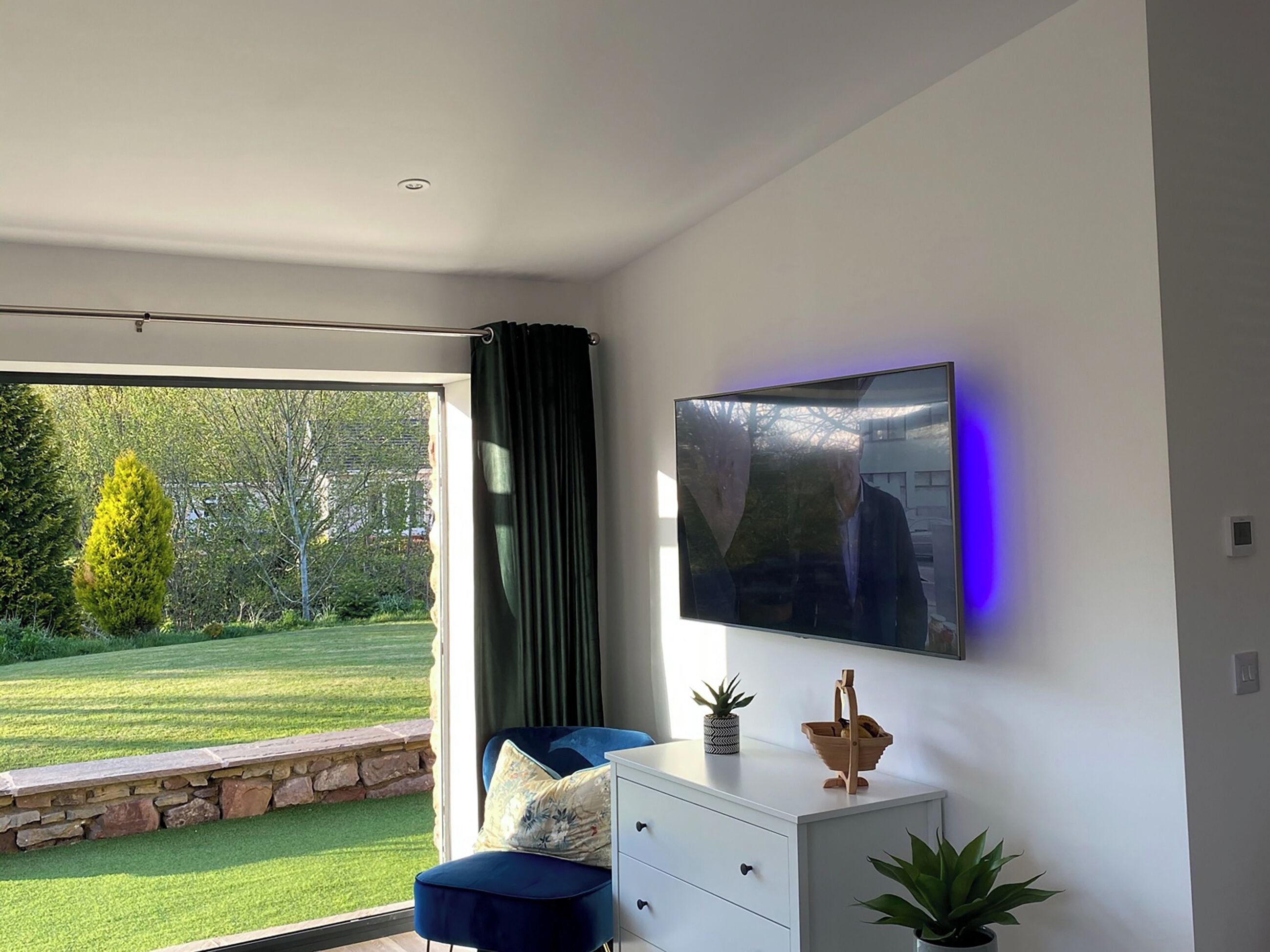 A modern living room with a large TV, a sofa, a stone wall, and a view of a garden.
