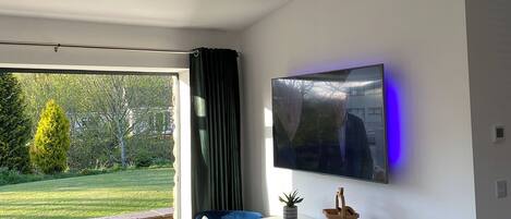 A modern living room with a large TV, a sofa, a stone wall, and a view of a garden.
