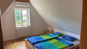 Un dormitorio con techo inclinado, una ventana, un radiador, una mesita de noche con lámpara y una cama con colcha colorida.