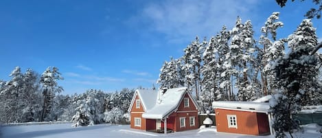 A snow-covered landscape with a red wooden house and a smaller structure, surrounded by snow-laden trees.