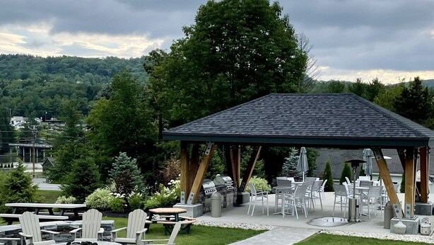 Un espace extérieur aménagé en terrasse, doté d’un sol carrelé en pierre, de chaises et de tables en bois, et entouré d’une pelouse verdoyante et d’arbres.