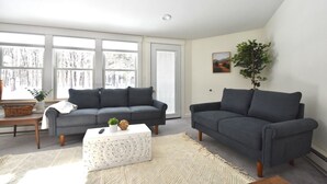 Een woonkamer met twee donkergrijze banken, een witte salontafel en een groot raam met uitzicht op bomen.