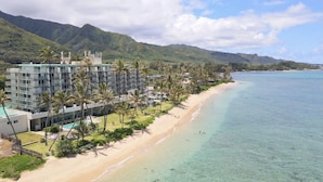 Un hotel frente a la playa con varios pisos, piscina y palmeras.