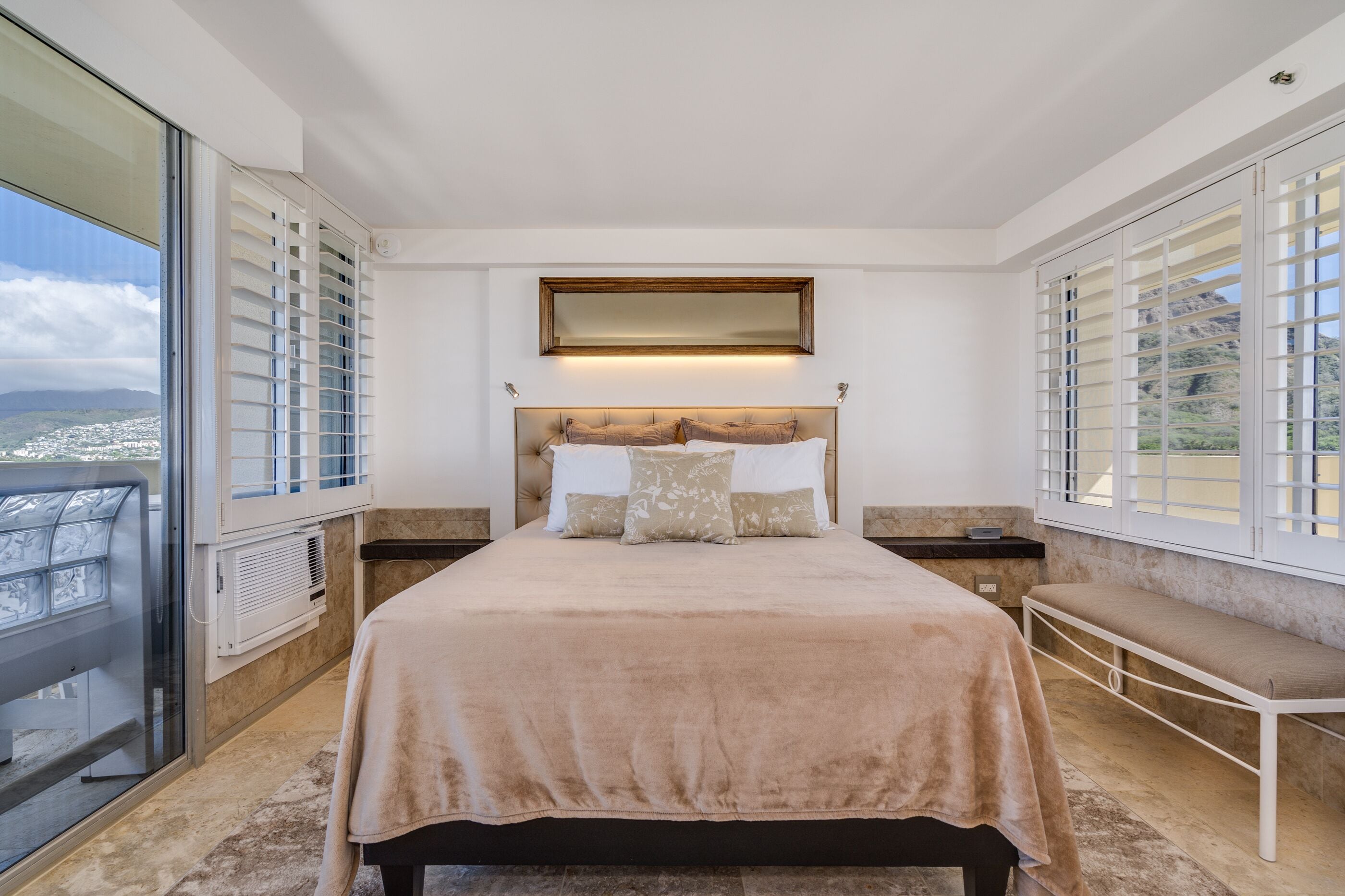 A bedroom with a large bed, a bench, a window with blinds, and a view of a cityscape.