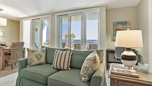 A green sofa with patterned cushions, a wooden side table with a lamp, and a view of the ocean through large windows. - Sunkissed Escape with spectacular views (Fernandina Beach)
