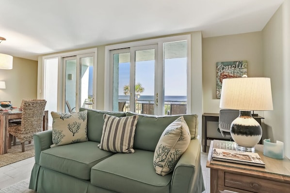 A green sofa with patterned cushions, a wooden side table with a lamp, and a view of the ocean through large windows.