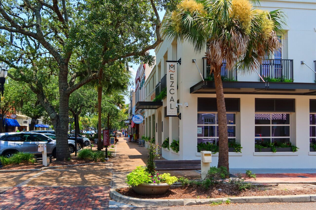 A street view with a building labeled "MEZCALI" featuring a covered front porch, palm trees, and a brick sidewalk.