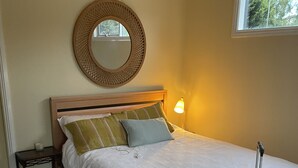 A neatly made bed with a headboard, a bedside lamp, a round mirror, and a suitcase on the floor. - Kestrel Place (Ocean Grove)