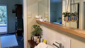 A bathroom with a large mirror, a wooden shelf with towels, a potted plant, and a sink with a chrome faucet. - Kestrel Place (Ocean Grove)